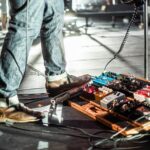 Guitarist in brown shoes stands beside a pedalboard onstage