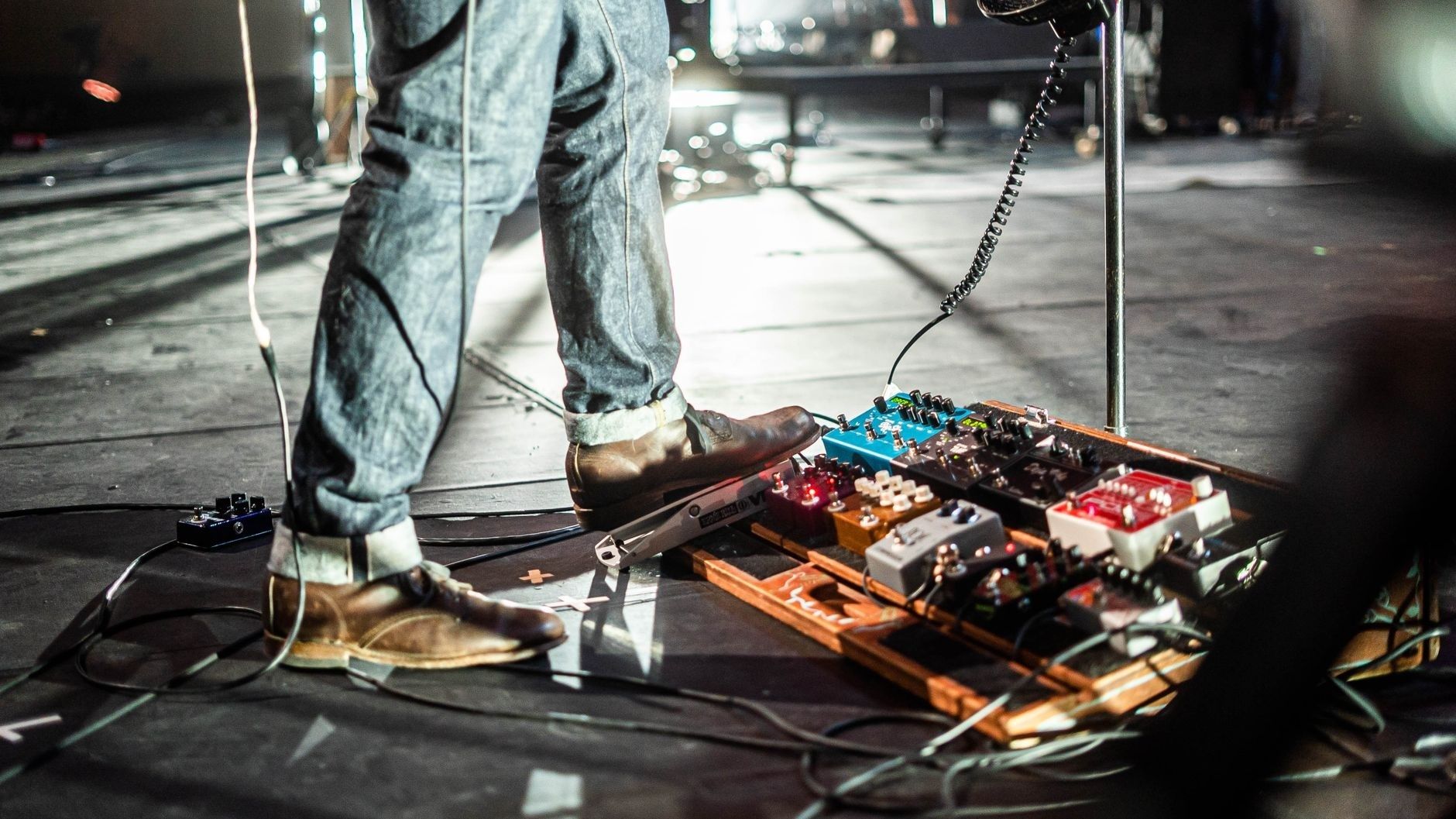 Guitarist in brown shoes stands beside a pedalboard onstage